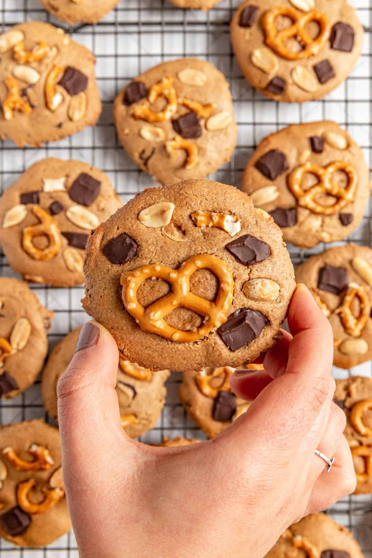 A person holding a Vegan Kitchen Sink Cookie over a rack of cooling cookies.