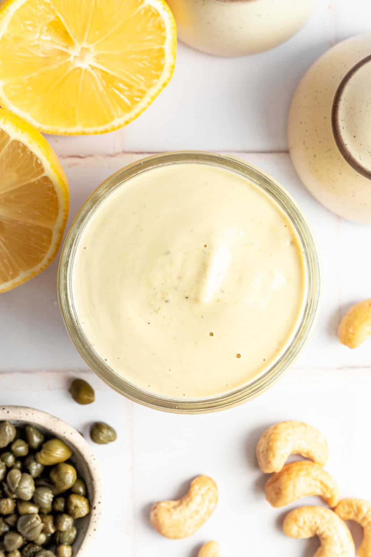 A jar of Vegan Oil-Free Cashew Caesar Salad Dressing surrounded by lemons, capers, and cashews.