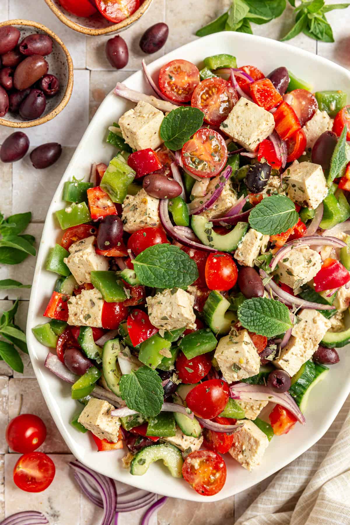 A platter of Vegan Greek Salad surrounded by tomatoes, olives, and oregano.