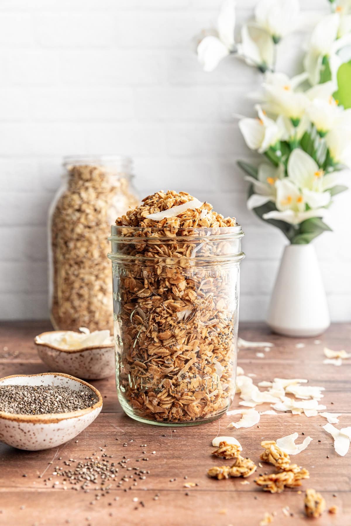 A jar of Easy Vanilla Coconut Granola with a jar of oats and a vase of flowers.