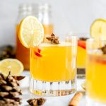 A glass of apple hot toddy, garnished with a lemon wedge, fresh apples, and star anise.