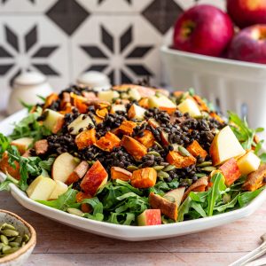 A platter of black lentil salad with a bowl of pepitas and a basket of apples.