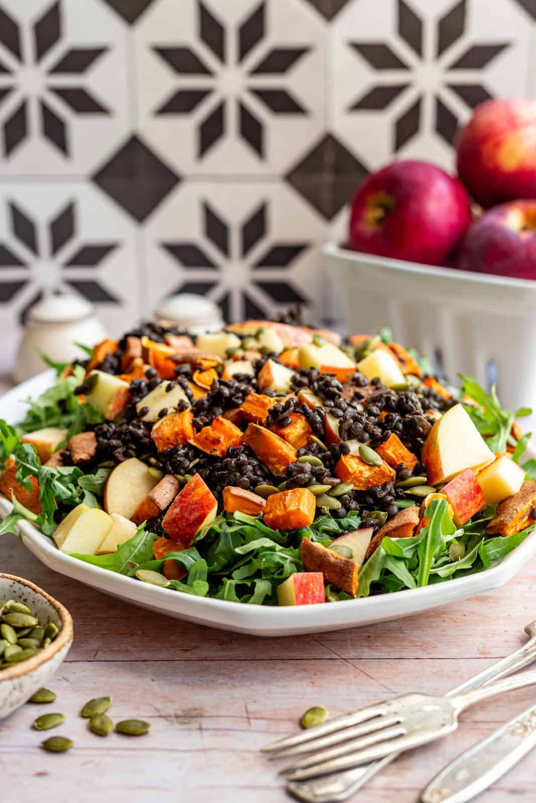 A platter of black lentil salad with a bowl of pepitas and a basket of apples.