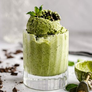 A glass with several scoops of vegan no churn mint chocolate chip ice cream, garnished with a sprig of mint.