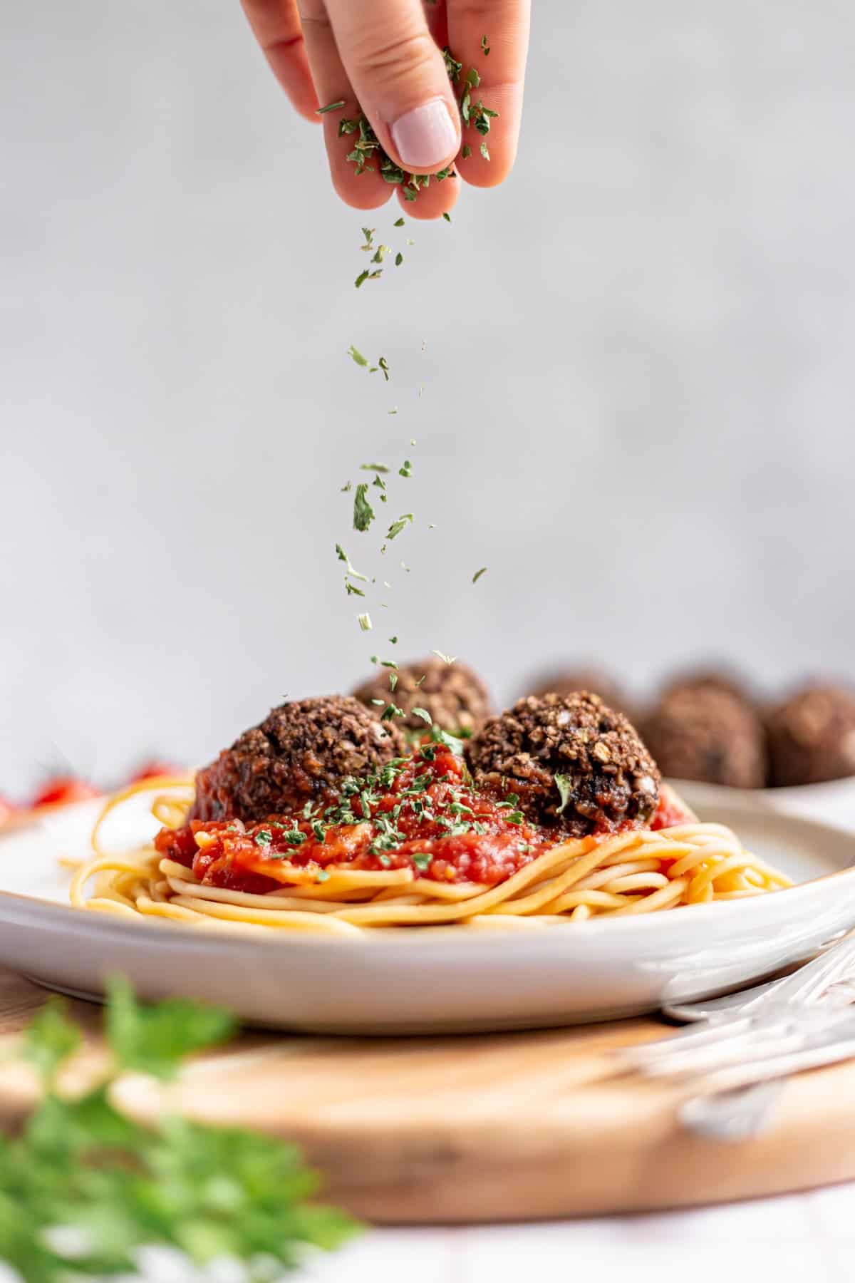 A person sprinkling herbs on a plate of vegan spaghetti and meatballs.
