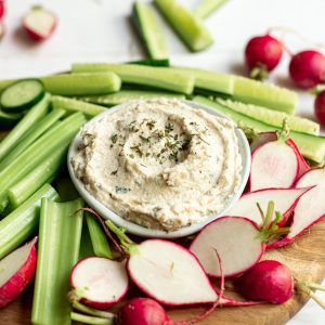 A bowl of vegan sour cream and onion dip on a tray of celery, cucumbers, and radish.