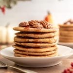 A stack of vegan sweet potato pancakes, topped with pecans and dripping with maple syrup.