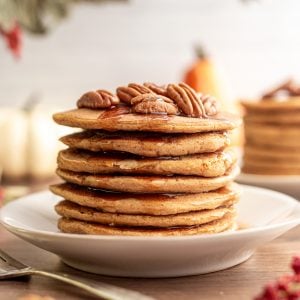 A stack of vegan sweet potato pancakes, topped with pecans and dripping with maple syrup.