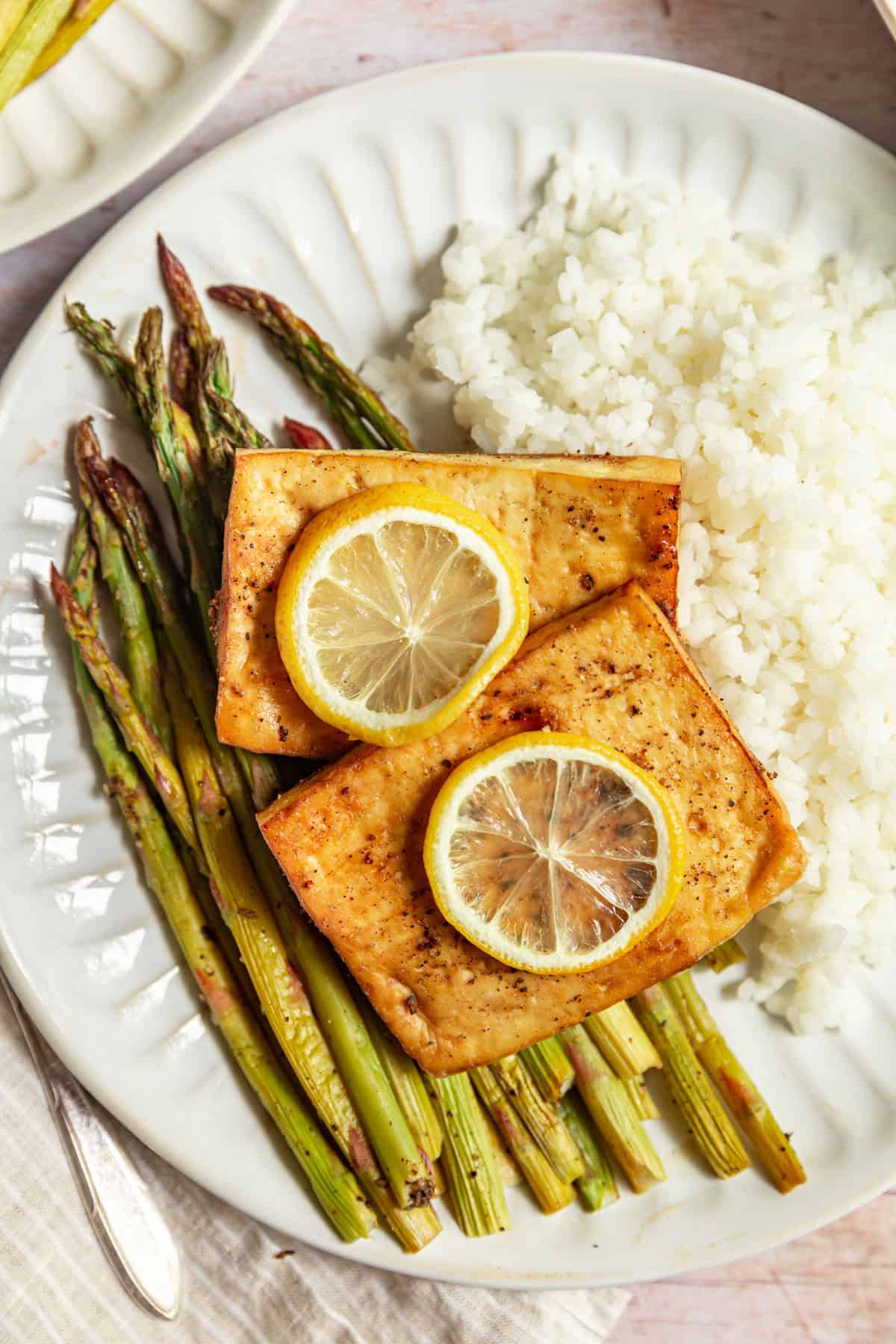 A plate of cooked asparagus, white rice, and lemon pepper tofu, topped with lemon slices.