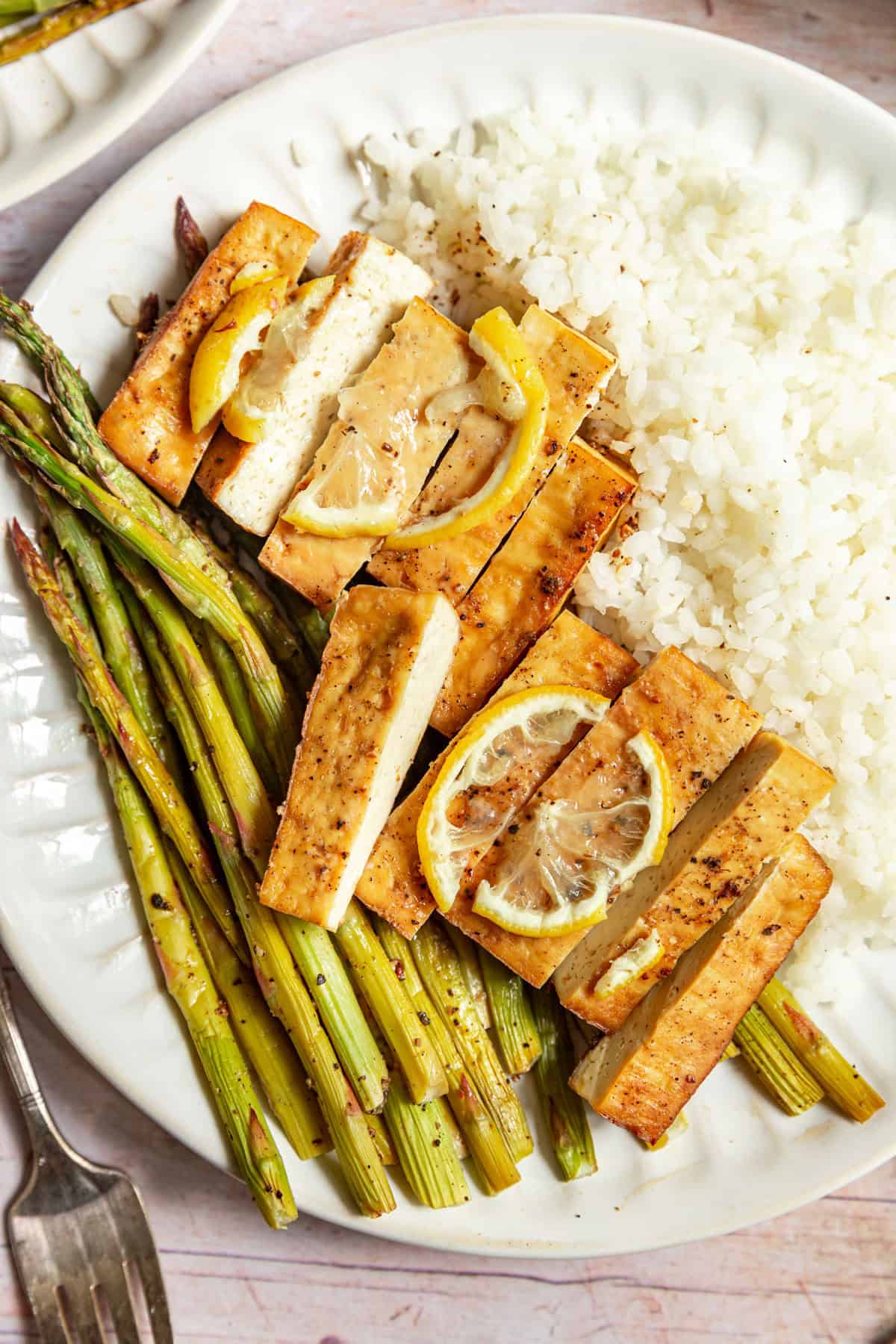 A plate of lemon pepper tofu sliced over rice and asparagus.