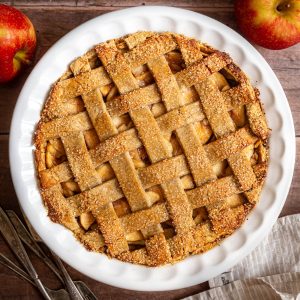 A freshly baked gluten free vegan apple pie, with forks, napkin and apples surrounding the pie.