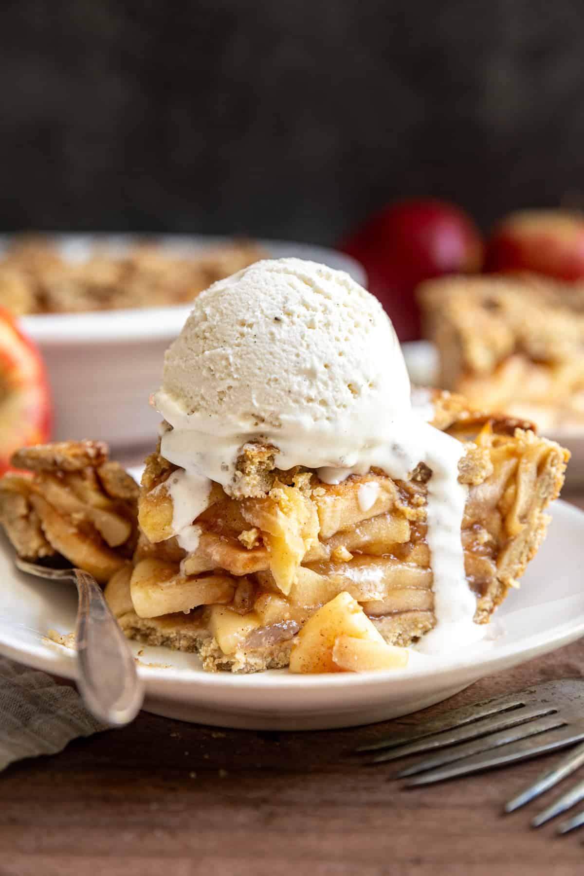 A slice of gluten free vegan apple pie on a plate, with a scoop of melting ice cream on top, and a bite taken out.