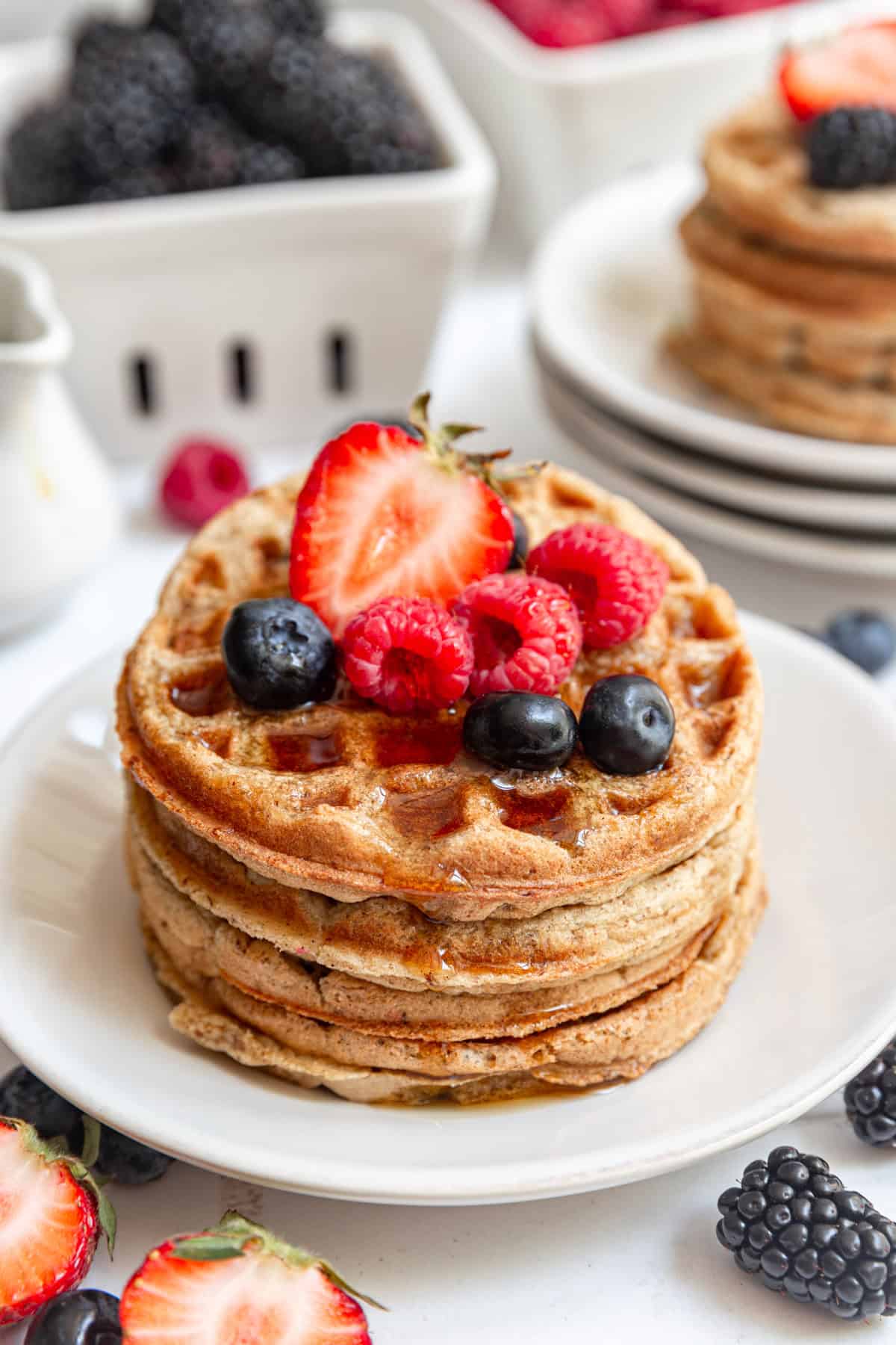 A stack of oat flour waffles topped with fresh berries and dripping with maple syrup.