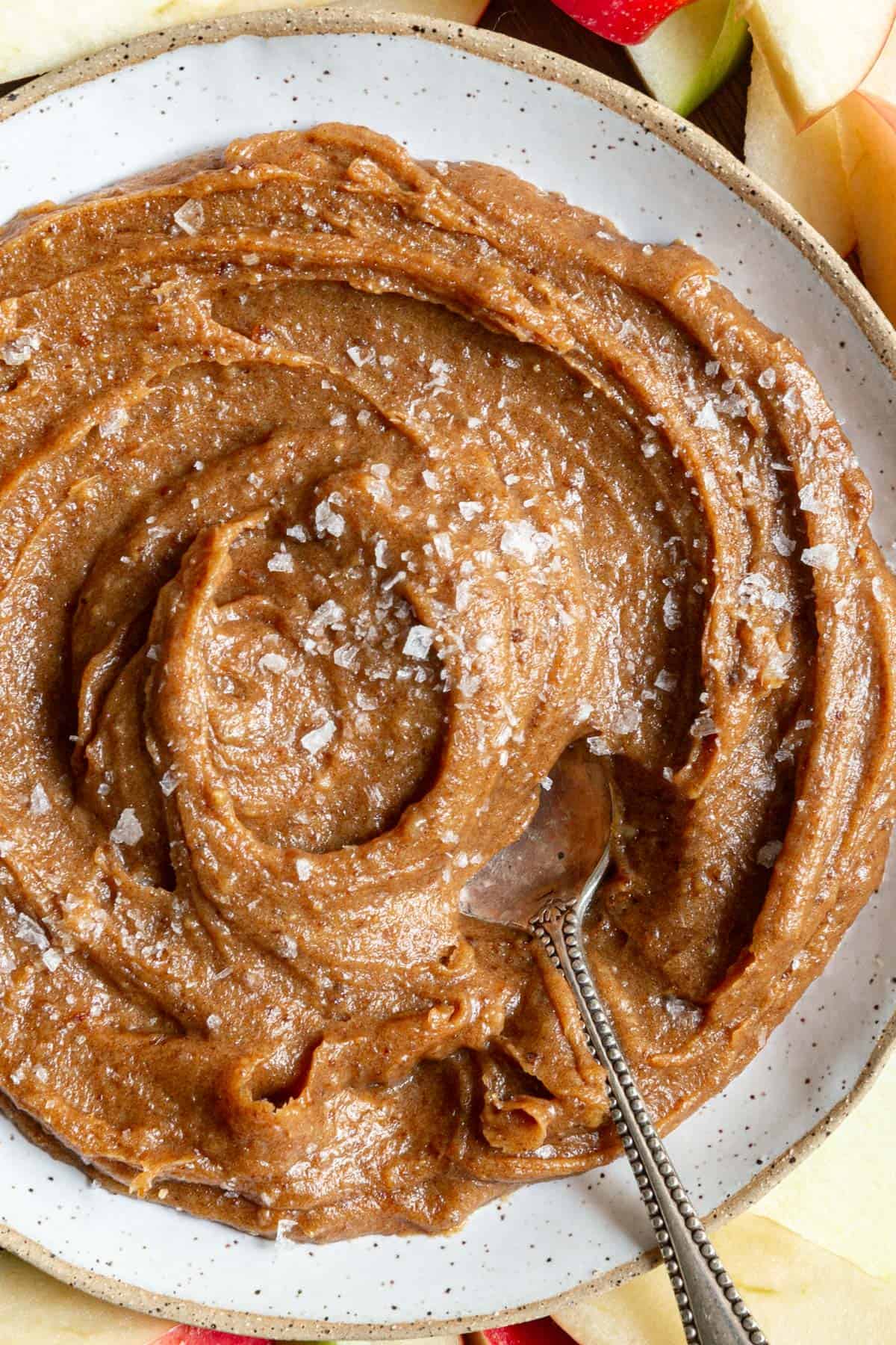 A bowl of Vegan Date Caramel Sauce topped with sea salt, with a spoon scooping some out.