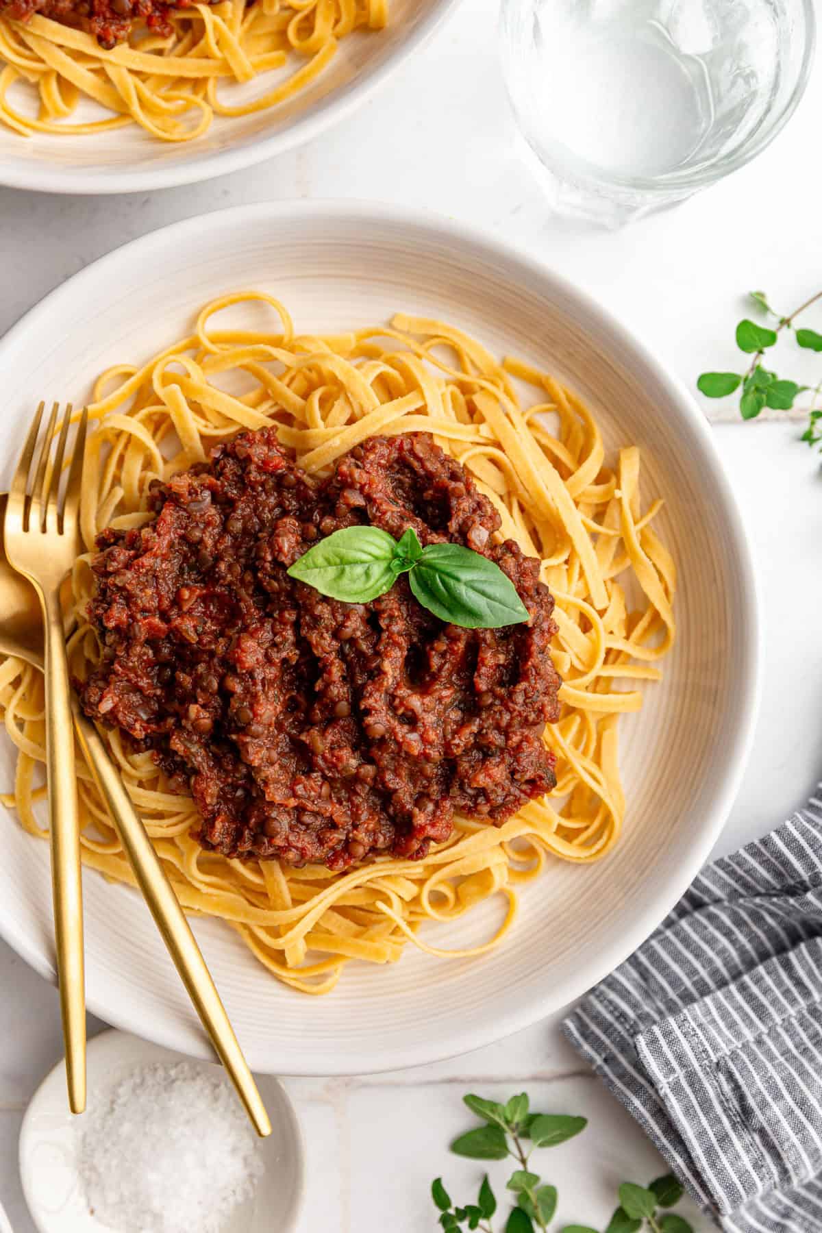 A bowl of lentil bolognese on top of linguine pasta, with a garnish of basil, and a fork and spoon in the bowl.