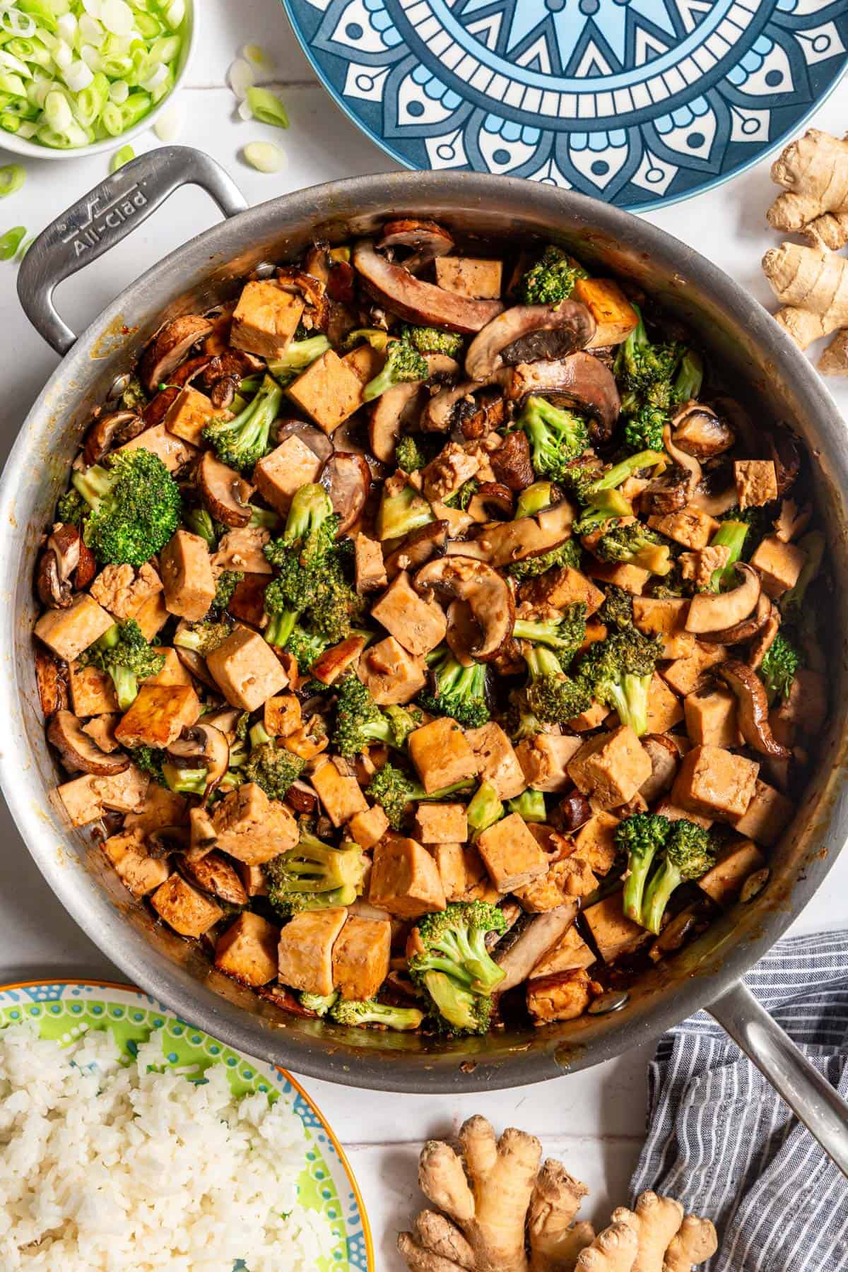 A pan of Tofu Broccoli Mushroom Stir Fry, with plates of rice and a bowl of scallions.