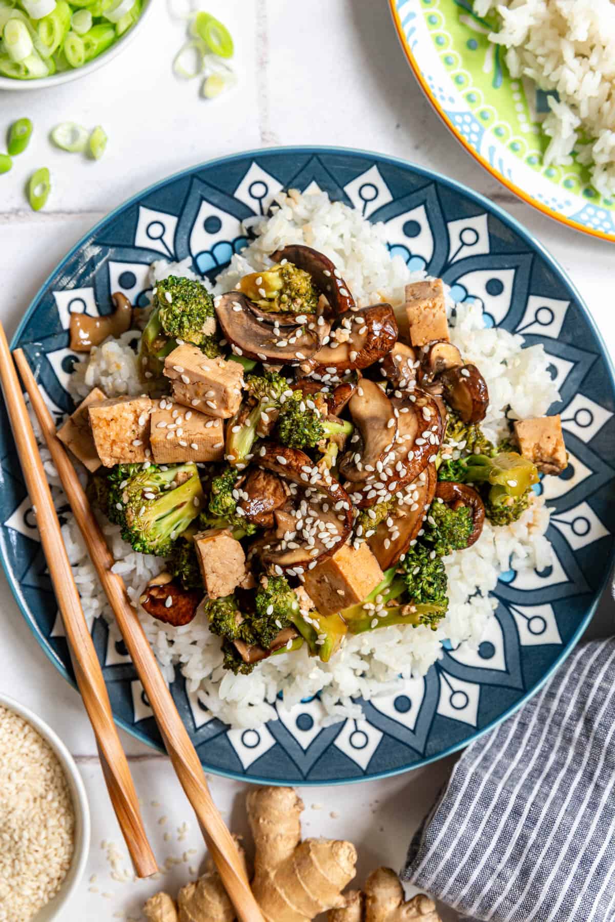 A plate of Tofu Broccoli Mushroom Stir Fry served over rice, with chopsticks.