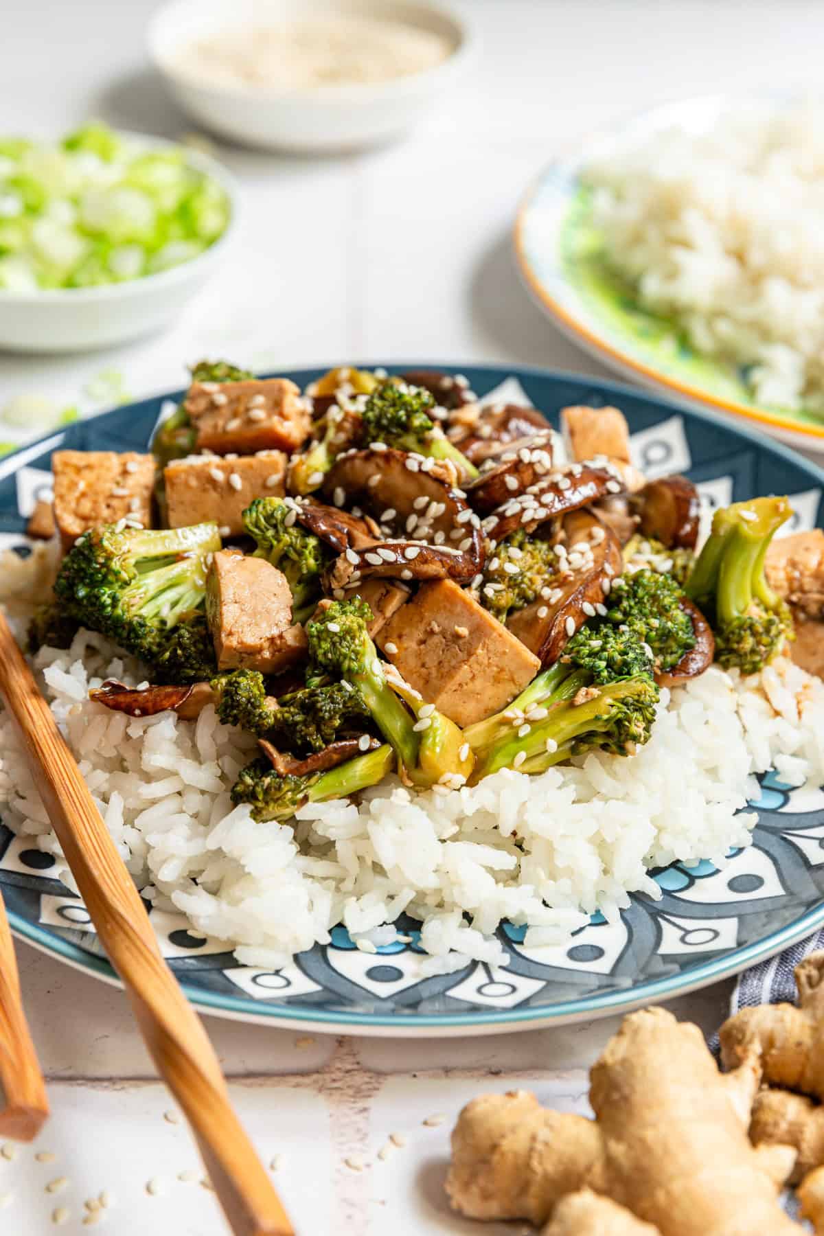 A plate of Tofu Broccoli Mushroom Stir Fry with bowls of rice, sesame seeds, scallions, and ginger.