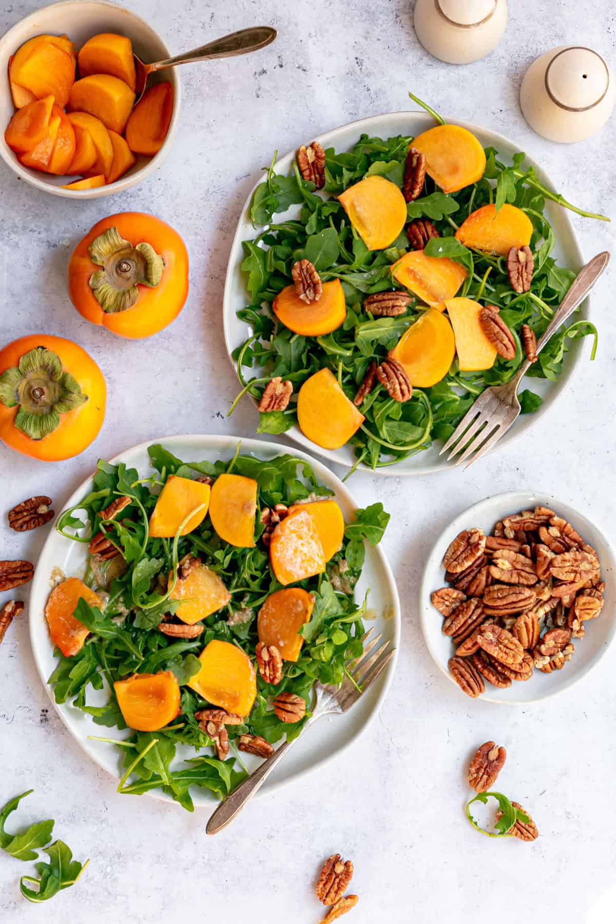Two plates of arugula persiommon salad, surrounded by whole persimmons, a bowl of sliced persimmons, and pecans.