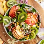 A bowl of kiwi apple spinach salad on a board, surrounded by ingredients used in the salad.
