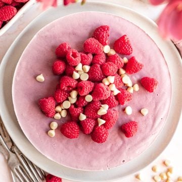 A plate with a vegan raspberry cheesecake, decorated with fresh raspberries and white chocolate chips.