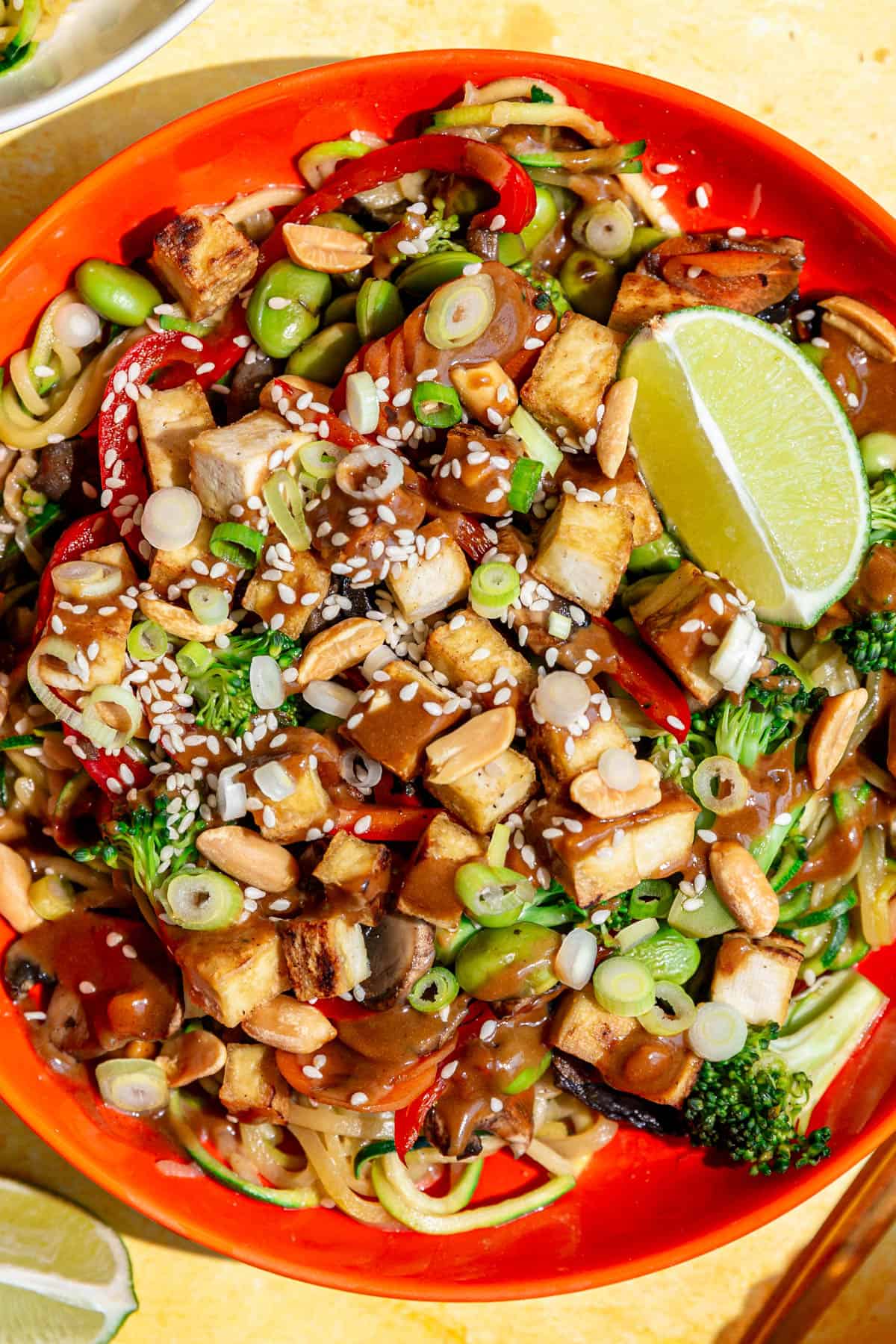 A bowl of zoodle pad Thai, drizzled with peanut sauce, garnished with sesame seeds and a lime.