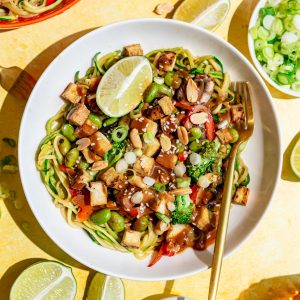 A bowl of zoodle pad thai, garnished with a lime wedge, with a fork, ready to eat.