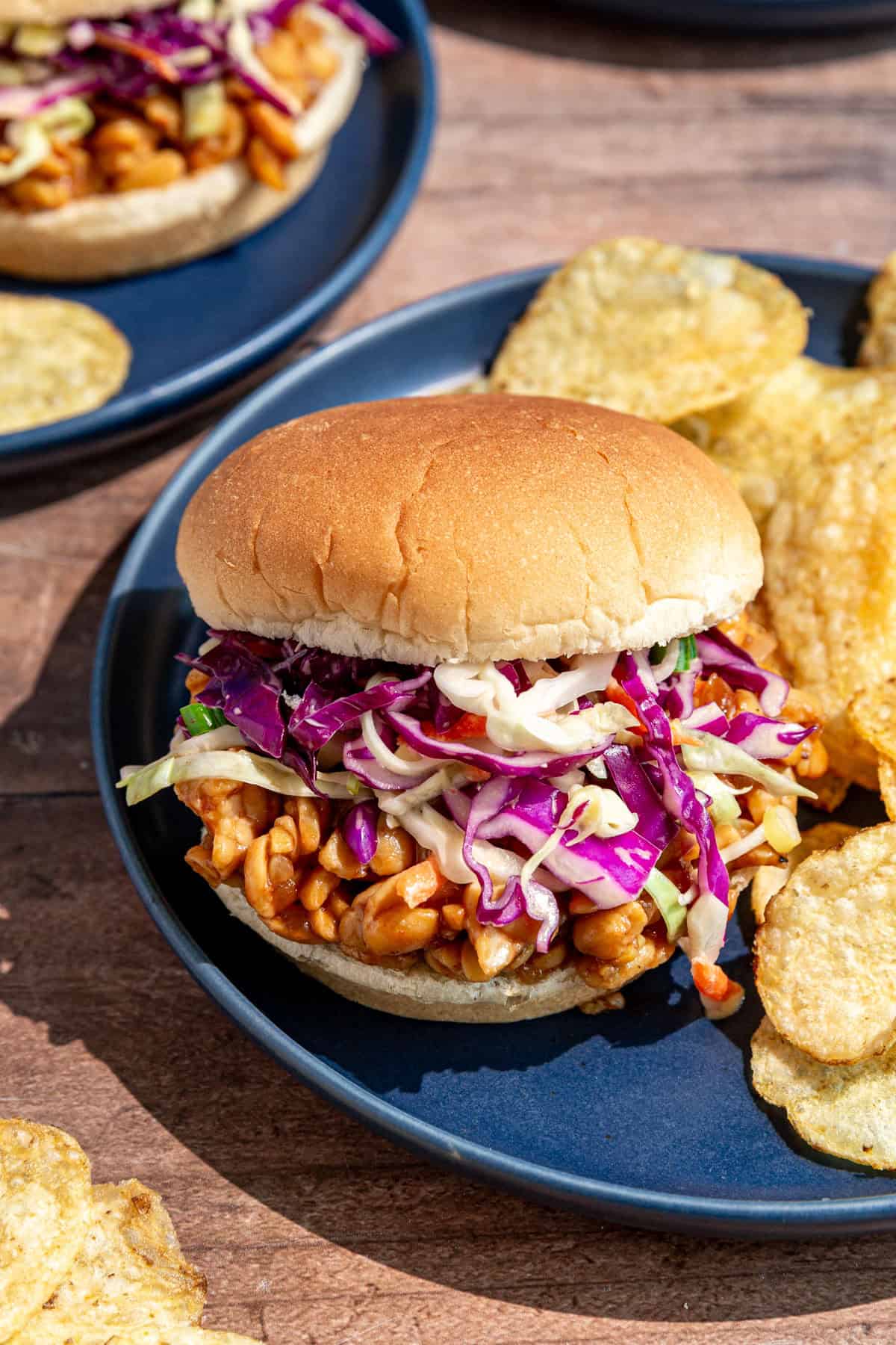 A bbq tempeh sandwich on a plate with chips.