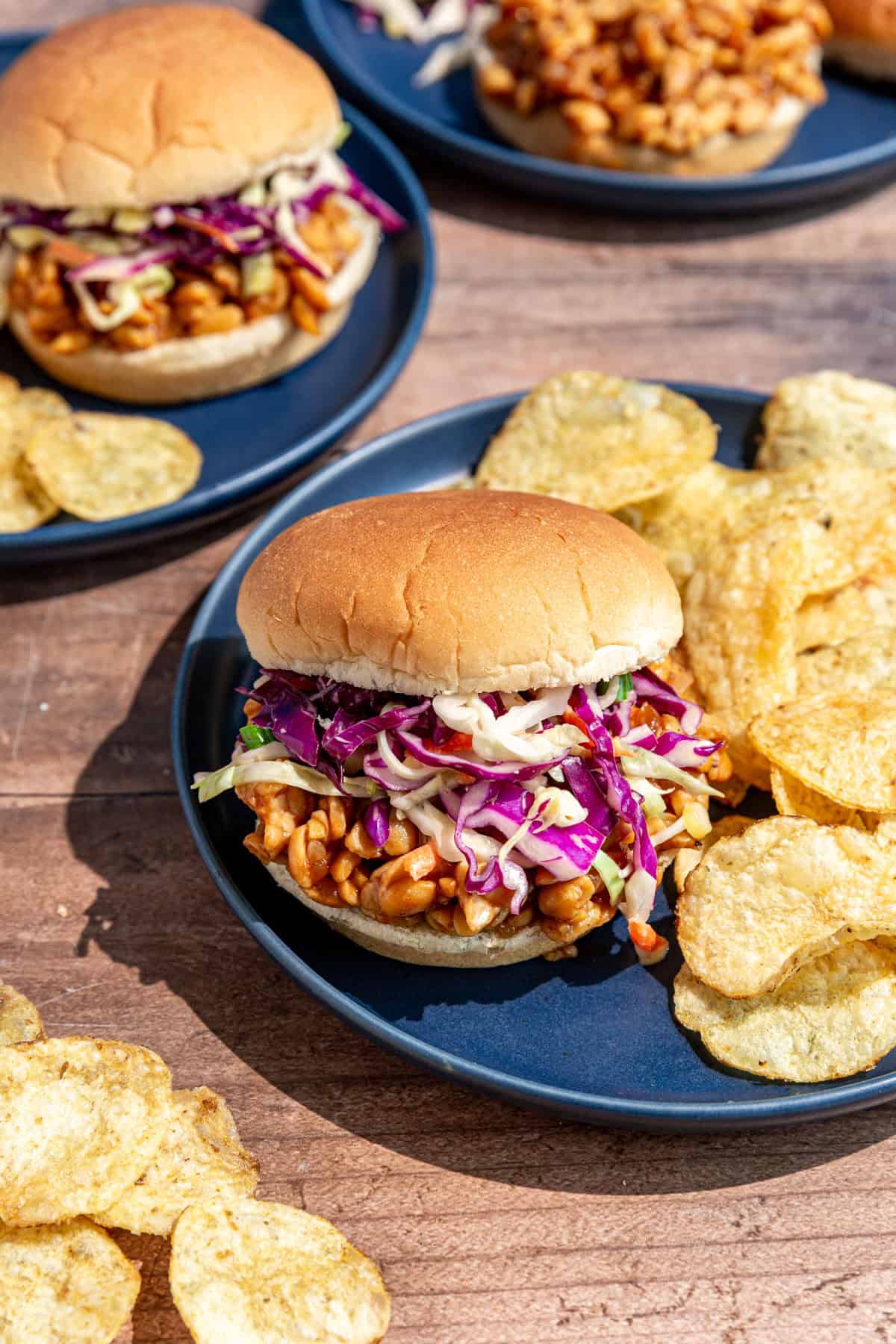 A bbq tempeh sandwich on a plate with chips.