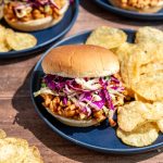 A bbq tempeh sandwich on a plate with chips.