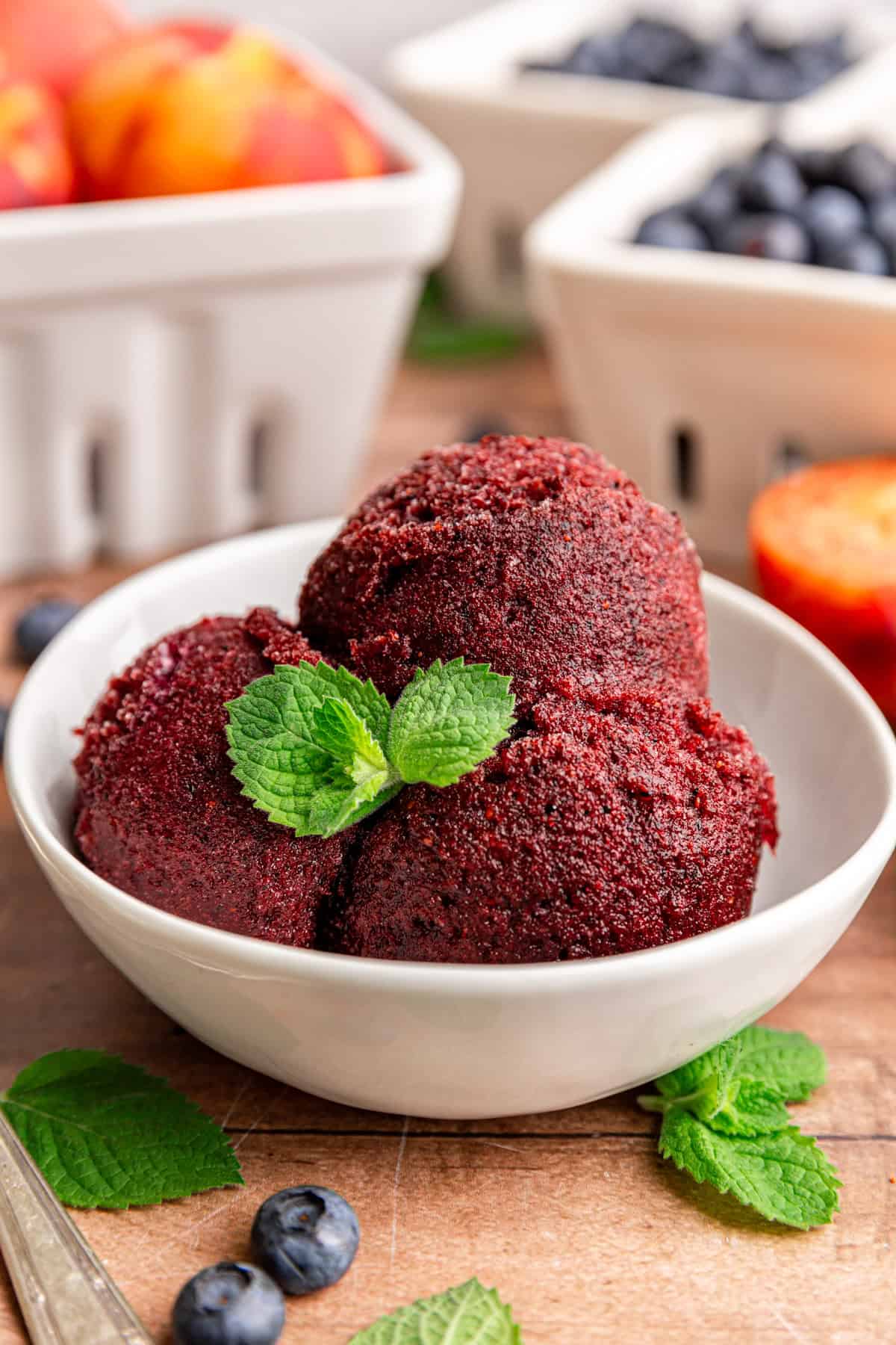 A bowl of Blueberry Peach Sorbet, garnished with mint, with a few mint leaves and berries surrounding it.