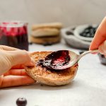 A person spreading sugar free blueberry jam onto an english muffin.