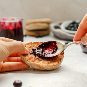 A person spreading sugar free blueberry jam onto an english muffin.