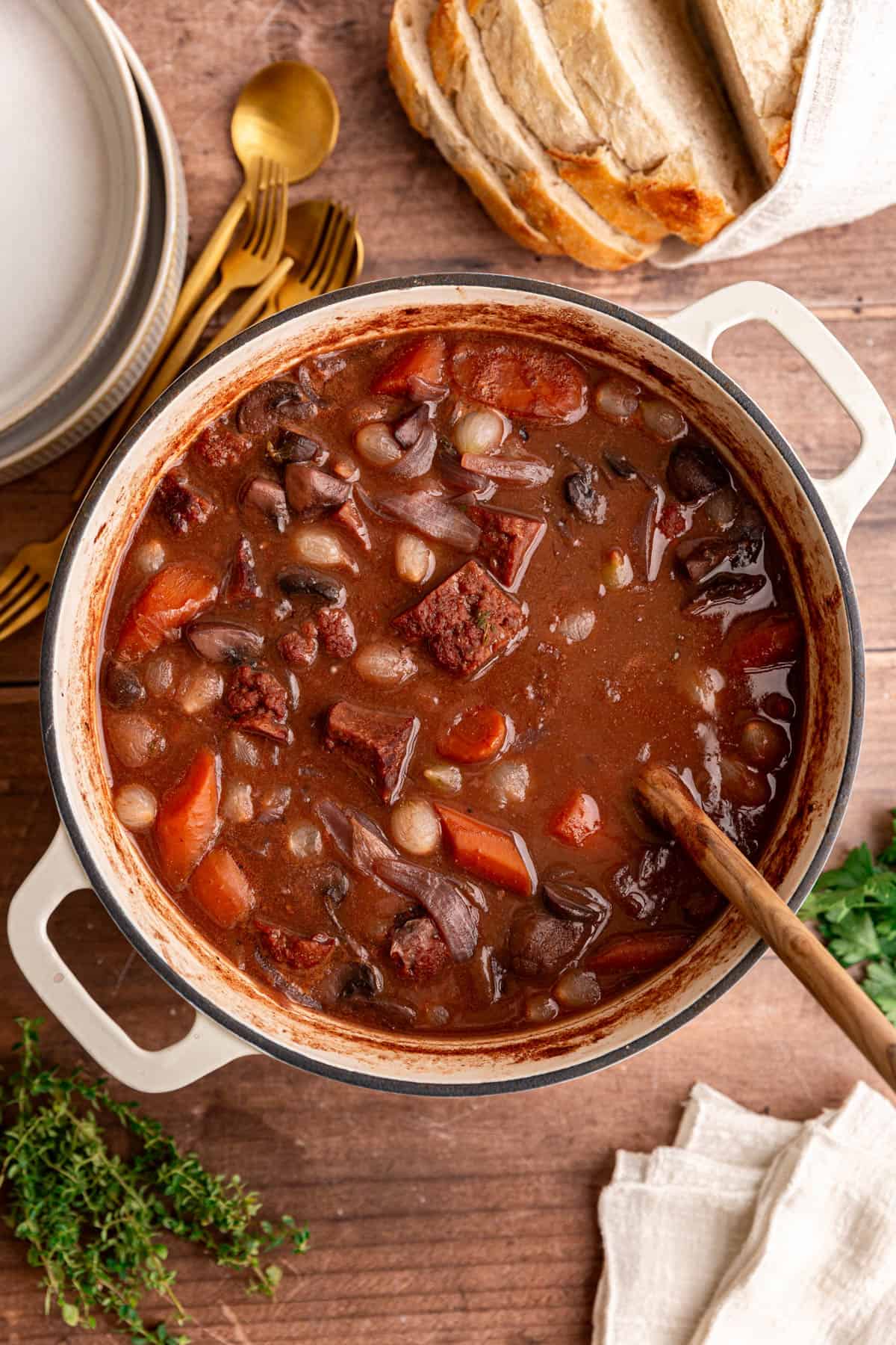 A pot of vegan beef bourguignon, freshly cooked, with empty bowls and garnishes surrounding it.