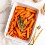 A baking dish of vegan glazed carrots, garnished with a large bunch of thyme, along with several plates, forks, and glasses.