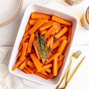 A baking dish of vegan glazed carrots, garnished with a large bunch of thyme, along with several plates, forks, and glasses.