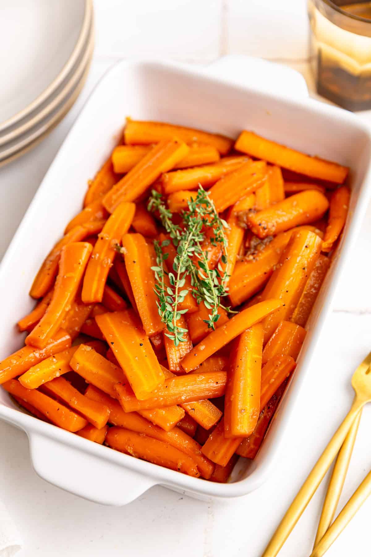 A baking dish of vegan glazed carrots, garnished with a large bunch of thyme, along with several plates, forks, and glasses.