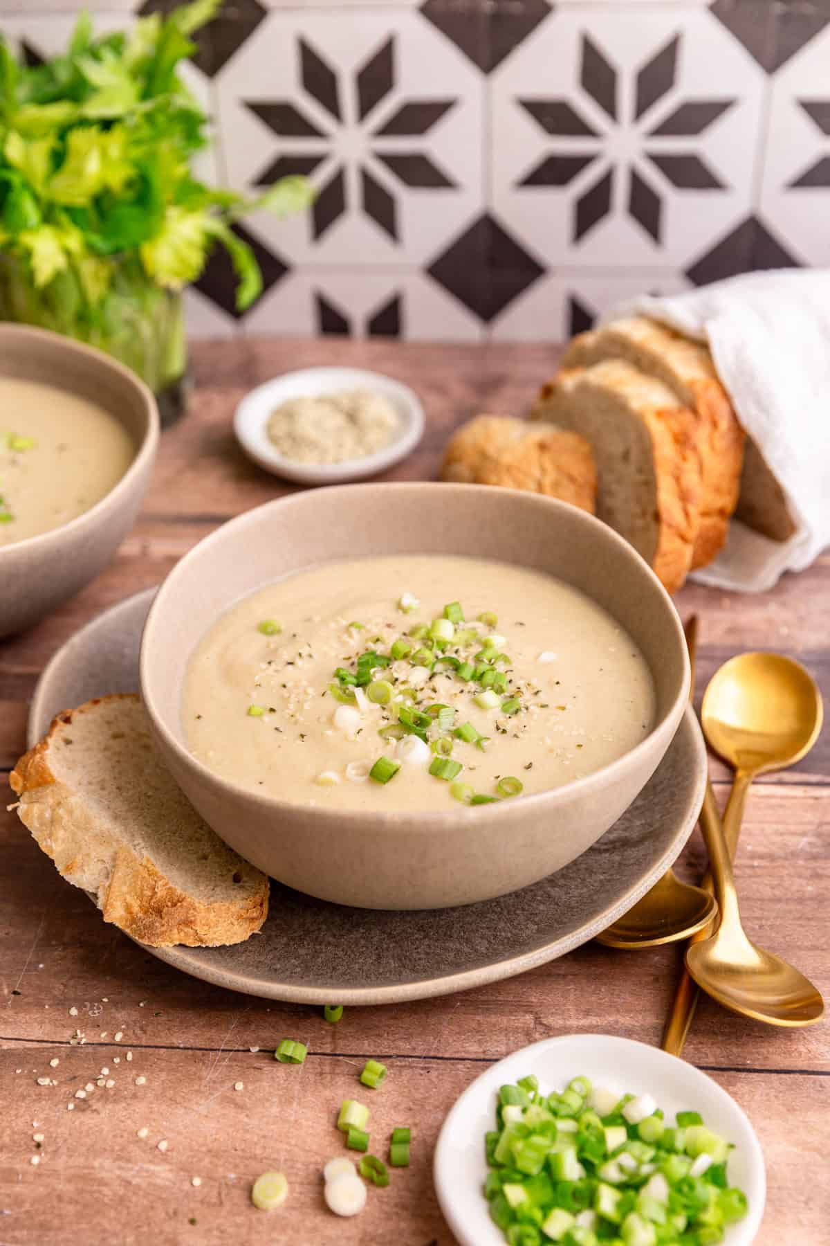 A bowl of vegan celery soup, on a table, with a loaf of bread and bowls of garnishes.