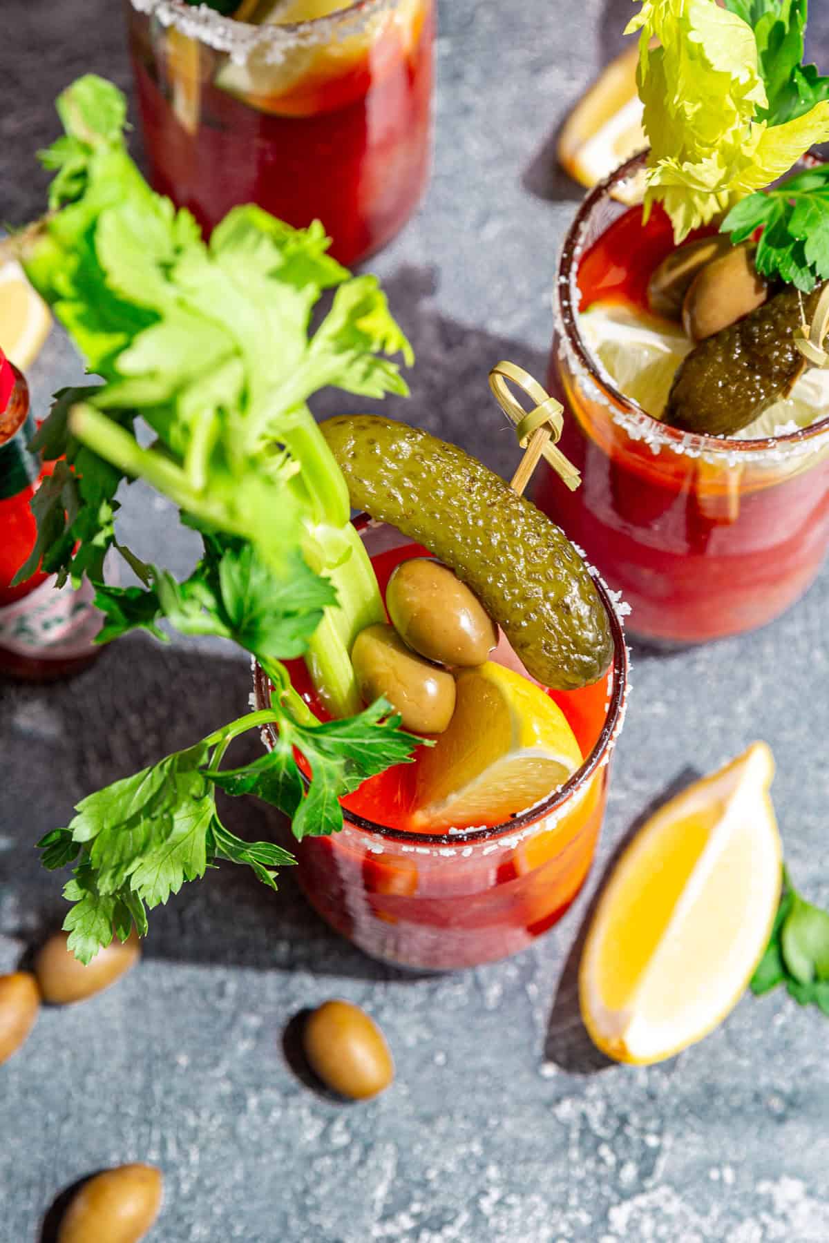 A top-down view of a bloody mary mocktail, garnished with celery, parsley, olives, pickles, and a lemon wedge.