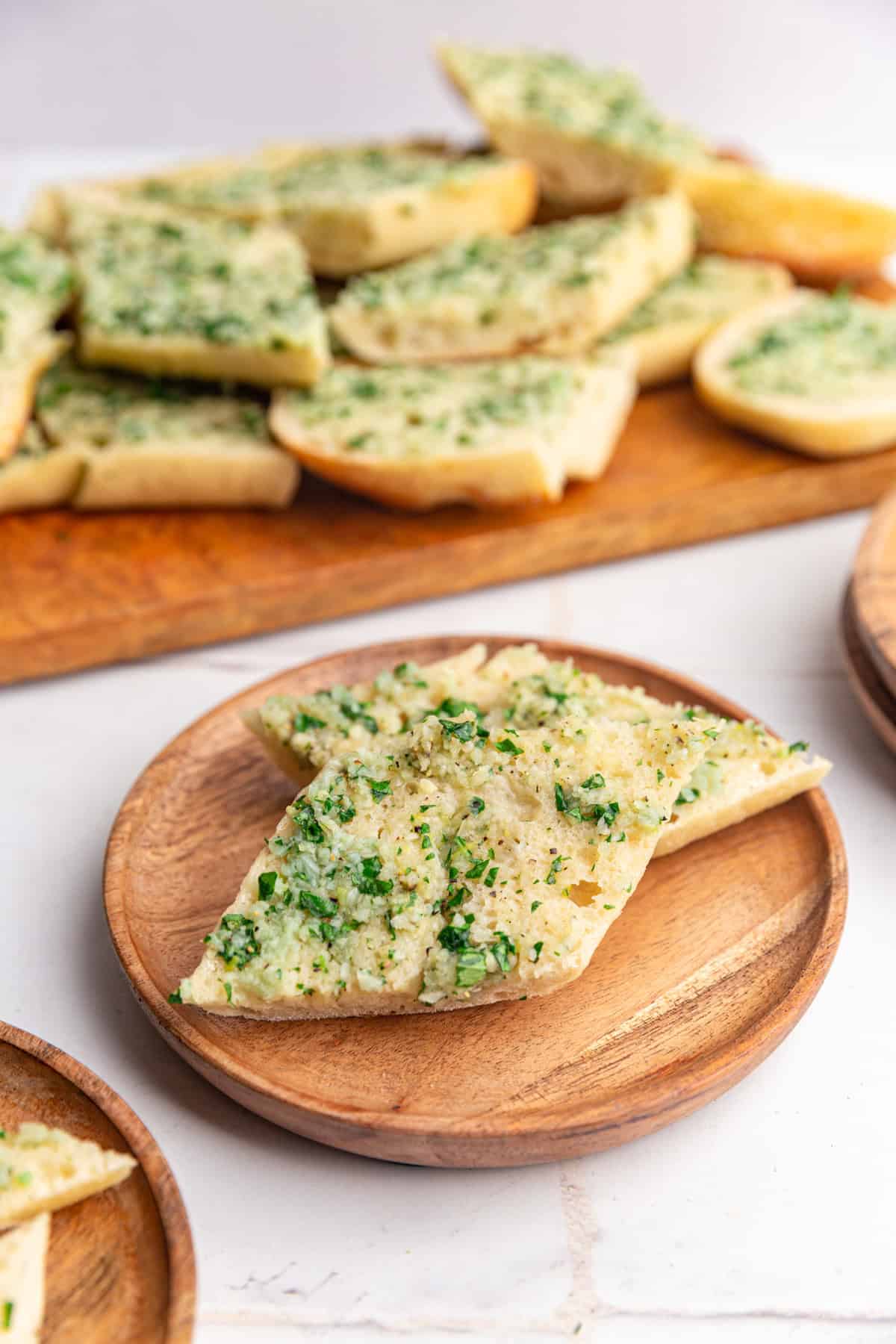 A plate with two slices of vegan garlic bread, and a tray of more garlic bread behind it.