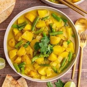 A bowl of green bean potato curry, with pita, cilantro, and lime wedges.