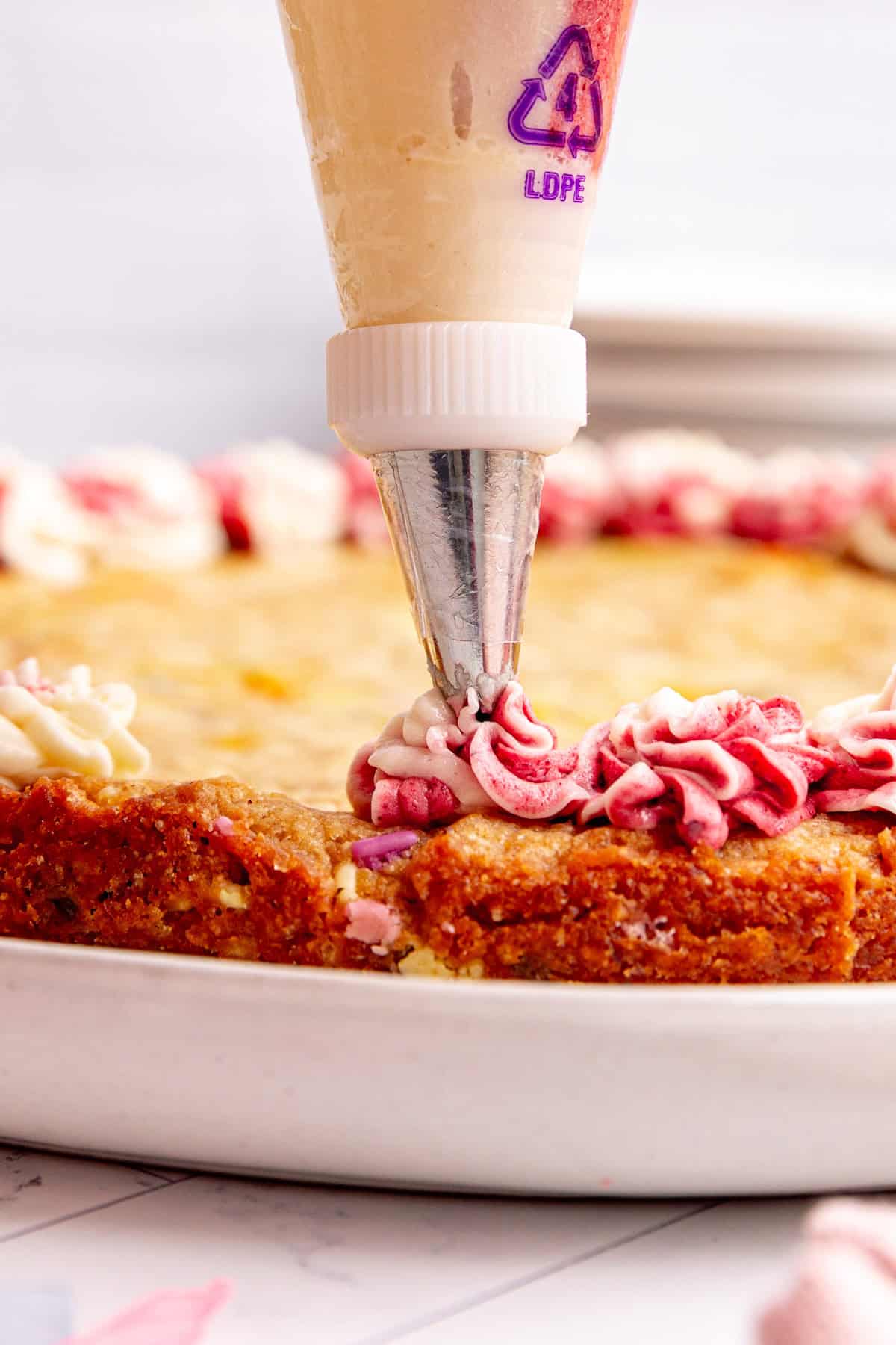 A piping bag frosting a dollop of pink and white frosting onto the edge of the cookie cake.
