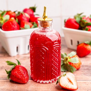 A bitters jar full of strawberry simple syrup, surrounded by fresh strawberries.