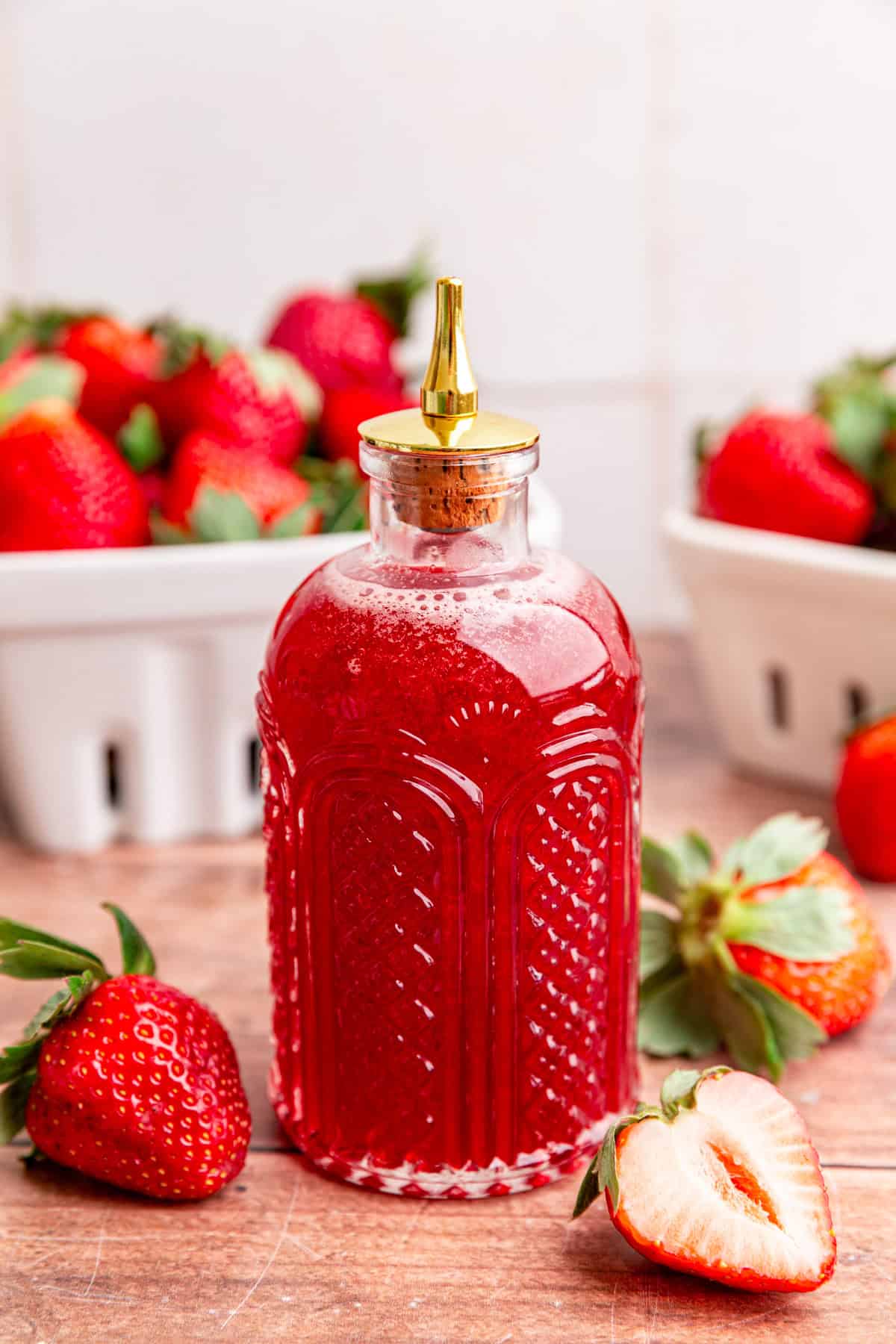 A bitters jar full of strawberry simple syrup, surrounded by fresh strawberries.