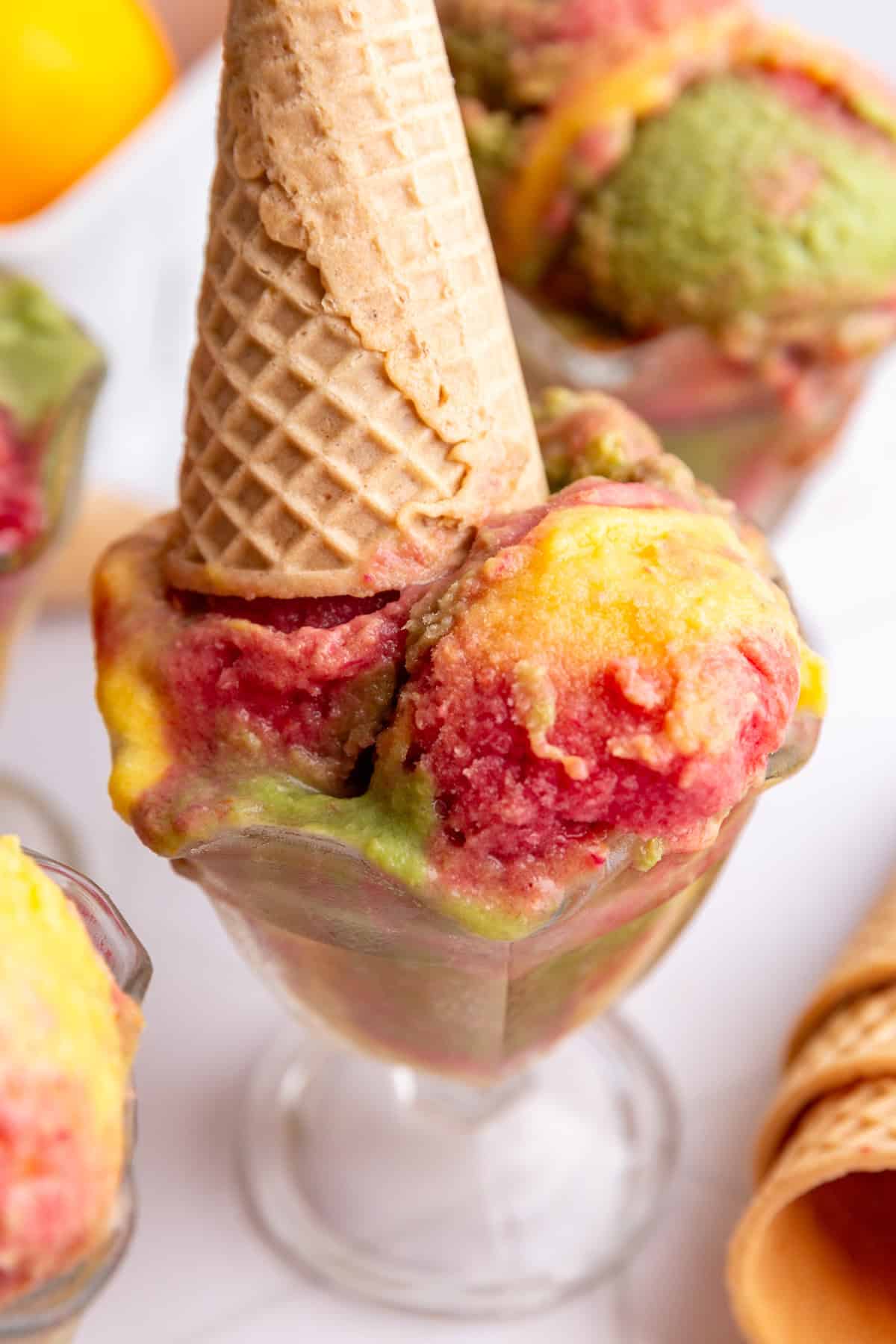 No churn dairy free orange, raspberry, and lime sherbet, mixed together in an ice cream glass, and an upside down cone.