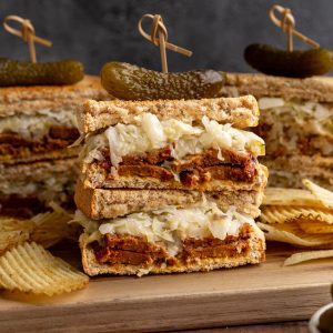 Several vegan pastrami sandwiches on a cutting board with potato chips.