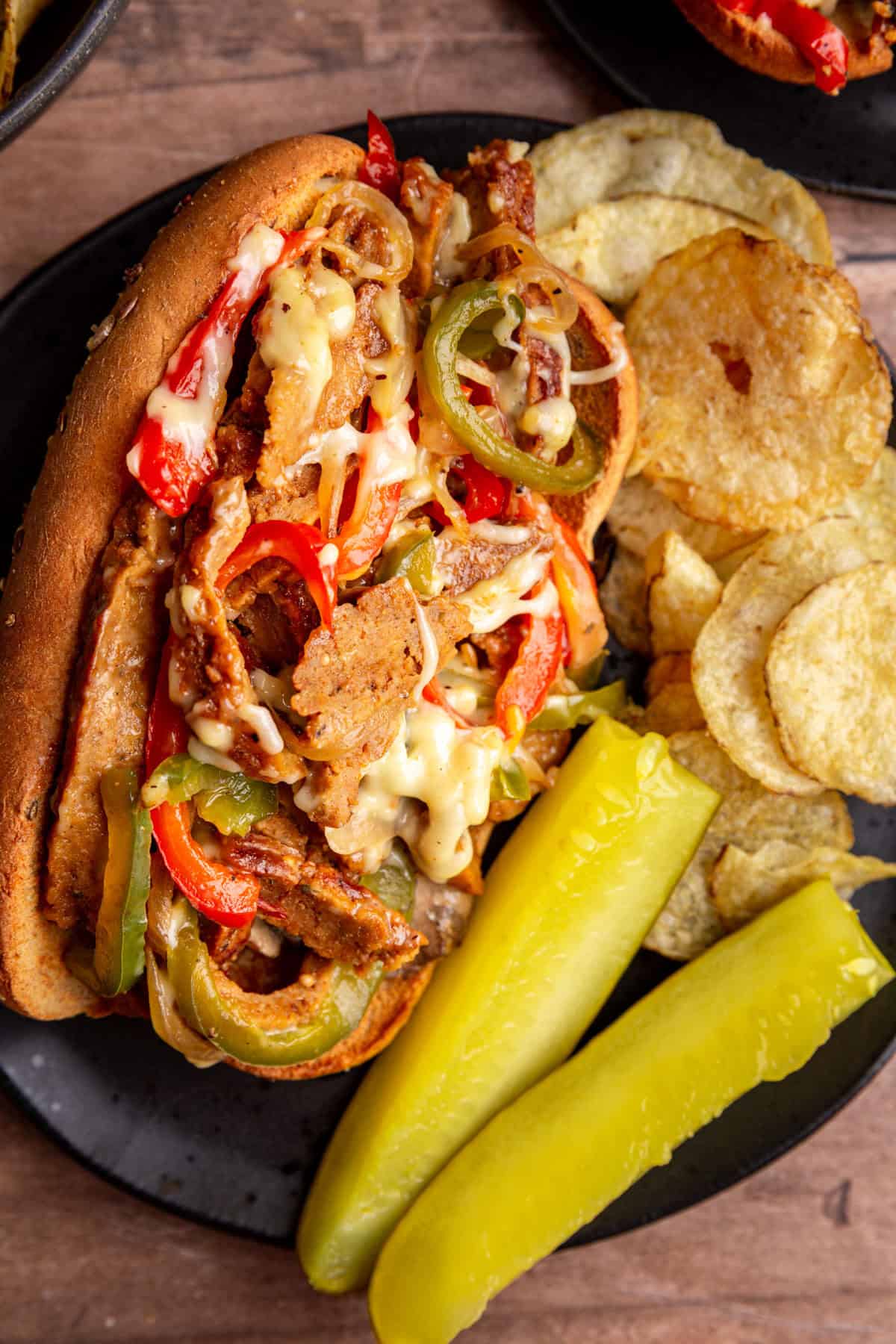 A plate with a vegan philly cheesesteak, chips, and pickle spears.