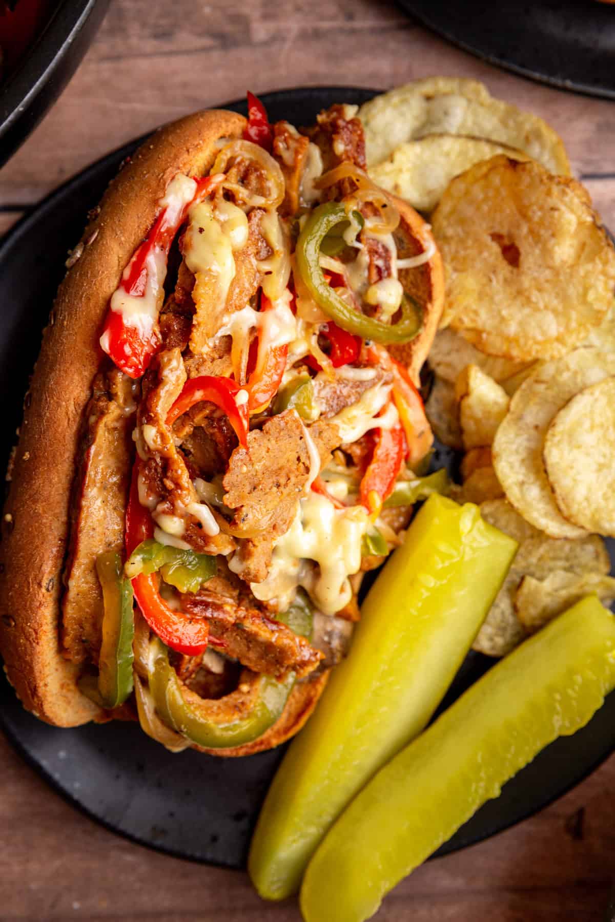 A plate with a vegan philly cheesesteak, chips, and pickle spears.