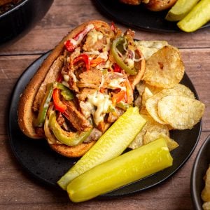 A plate with a vegan philly cheesesteak, chips, and pickle spears.