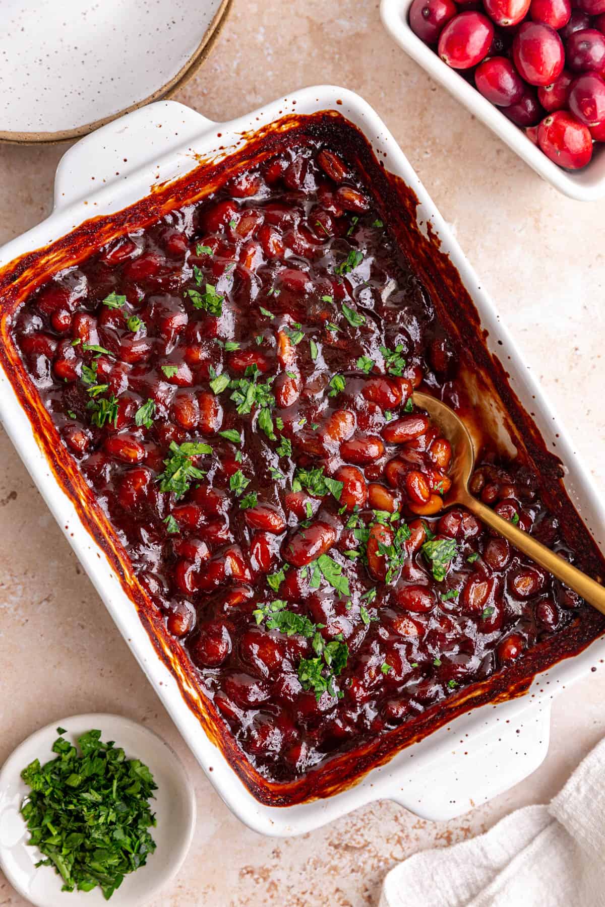 A dish of freshly baked cranberry bbq baked beans, garnished with parsley, with a spoon scooping some out.